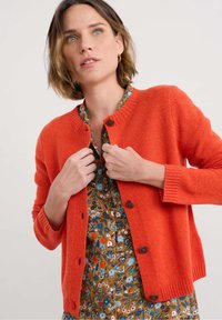 Cardigan en tricot orange à encolure ronde, avec des poignets côtelés et des boutons marron. Porté par-dessus une robe à motifs floraux.