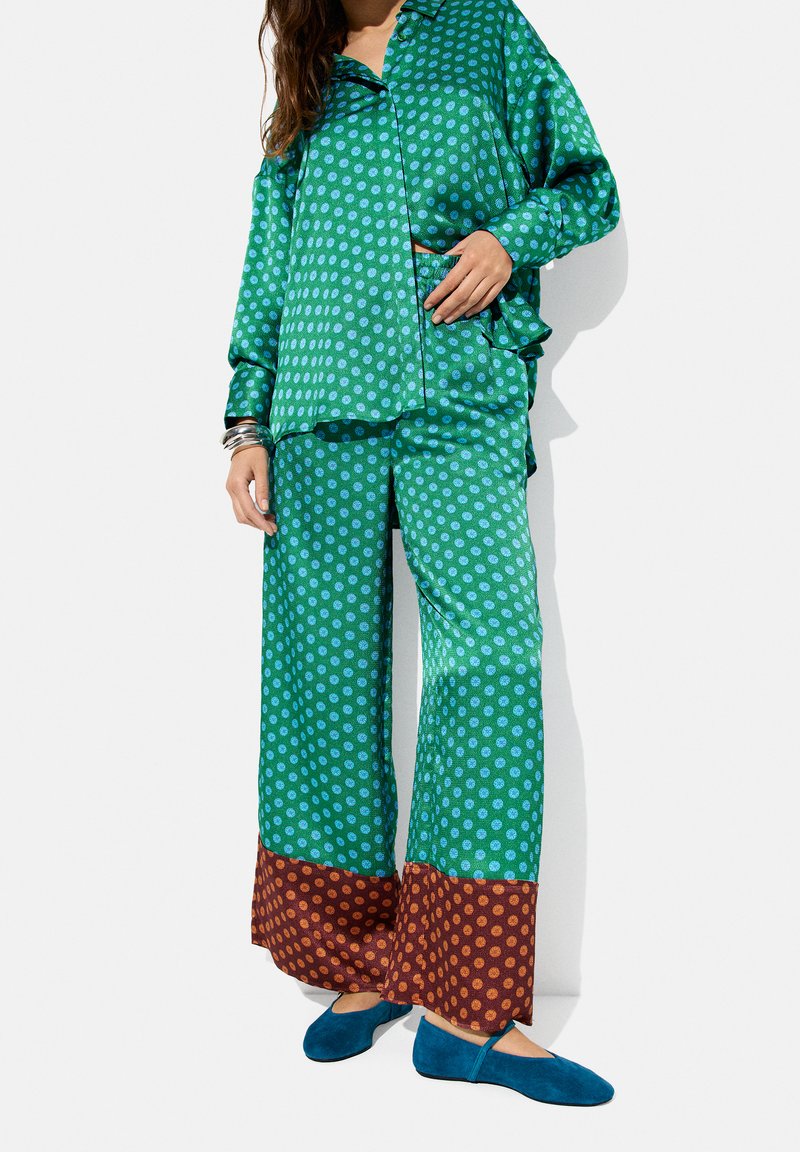 Zielony komplet piżamy w kropki z szerokimi spodniami, kontrastującymi brązowymi mankietami i niebieskimi akcentami. Miękka tkanina o satynowym wykończeniu. Niebieskie kapcie.