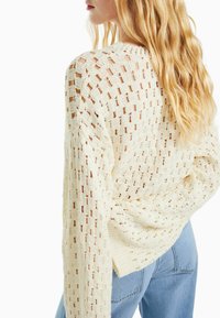 Stickad tröja i ljuskräm med ett öppet vävmönster, långa ärmar och en avslappnad passform. Texturen är mjuk med ribbade detaljer vid muddarna.