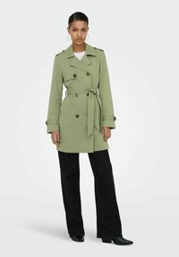 Femme portant un trench-coat vert clair ceinturé, pantalon large noir, bottes noires à bouts pointus, debout devant un fond clair uni.