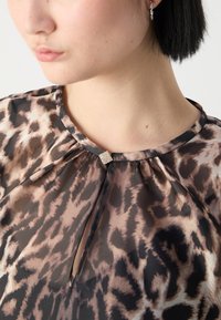 Blouse en transparence, marron, avec un motif léopard. Elle présente un col plissé et une petite décoration carrée au niveau du col.