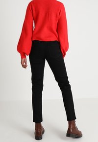 Punainen neulepusero, jossa on ilmapallohihat, yhdistetty mustiin kapealahkeisiin housuihin ja ruskeisiin nauhallisiin saappaisiin. Yksinkertainen muotoilu ilman näkyviä kuvioita.