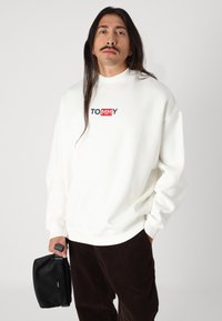 Bijela oversized dukserica s visokim ovratnikom, sa "TOMMY" logotipom u crvenoj i mornarskoj plavoj boji. Uparena s tamnosmešnim kašmir hlačama i crnom torbom.