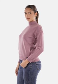 Donna con capelli scuri raccolti in una coda di cavallo, che indossa orecchini a cerchio d'oro, un maglione a spalle buttonato di color malva e jeans blu, in piedi di lato.