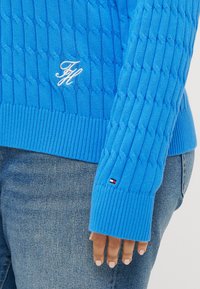 Pulover tricotat albastru cu un model de cablu, tiv și manșete nervurate, și un logo alb pe piept, cu un detaliu mic al logo-ului pe manșetă.