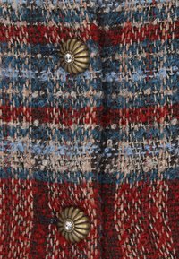 Vörös, kék és bézs színű szőtt textil, texturált mintával. A réz gomb, amelyen egy strasszkő díszítés található, jellegzetesen kiemelkedő.