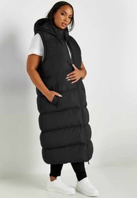 Svart pufferväst med huva, dragkedjestängning och sidofickor. Har en quiltad design med en lång silhuett och ribbad textur vid kanterna.