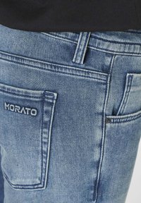 Svijetloplave traperice s vidljivim stražnjim džepom, s izvezenim imenom branda "MORATO." Klasičan dizajn s pet džepova, blago izblijedjele.