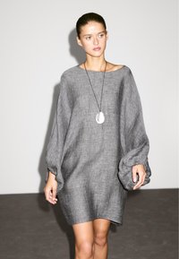 Abito in lino grigio con un design ampio e oversize, dotato di maniche larghe. Accentuato da una collana con pendente, presentato su uno sfondo neutro.