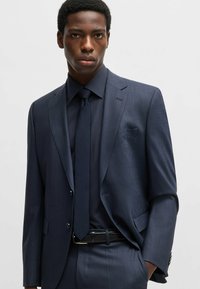 Completo su misura blu navy con un sottile motivo a quadretti, giacca a un bottone, cravatta abbinata e camicia a trama, completato da una cintura nera.