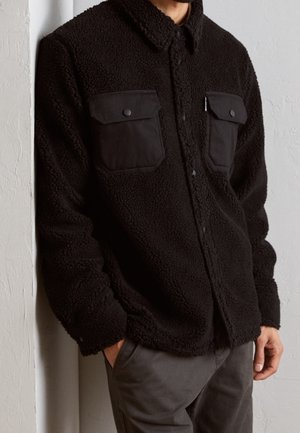 Mężczyzna w czarnej, puszystej kurtce zapinanej na guziki z dwoma przednimi kieszeniami z klapami, stoi z rękami w kieszeniach ciemnoszarych spodni na tle białej ściany.