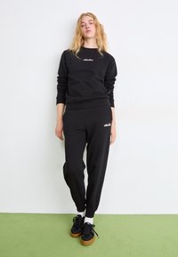 Sudadera negra y joggers confeccionados en tela suave, con puños acanalados, que presentan un logo bordado en el pecho y el muslo. Zapatillas negras con suelas de goma.