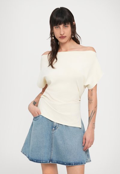 Εκτός ώμου κρέμ τοπ με σχεδιασμό σε στυλ drape, συνδυασμένο με ανοιχτό μπλε τζιν φούστα με φαρδύ σχήμα και τσέπες.