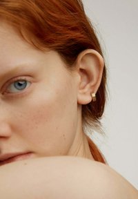 Pendiente de aro dorado con un diseño texturizado y estriado. Primer plano de la oreja de un modelo y parte de la cara, con piel clara y cabello rojo.