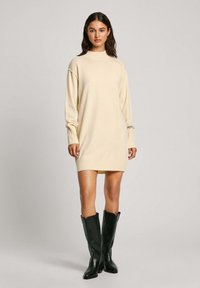 Abito maglione crema con collo alto, maniche lunghe, polsini e orlo a coste. Indossato con stivali neri in pelle fino al ginocchio.