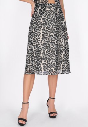 Kobieta ubrana w prześwitującą, sięgającą do połowy długość spódnicę w panterkę oraz czarne sandały na wysokim obcasie z paskiem wokół kostki, pozująca z jedną ręką na biodrze.