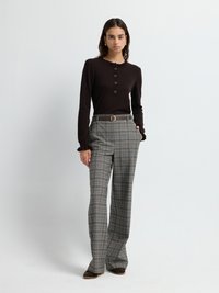 Maglietta a maniche lunghe marrone scuro con colletto sfrangiato e cinque bottoni dorati, abbinata a pantaloni larghi a quadri grigi e scarpe marroni.
