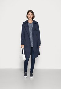 Casaco militar azul-marinho com botões duplamente abotoados, usado sobre uma blusa azul-marinho com padrão. Combinado com jeans slim, uma mala branca e ténis azul-marinho.