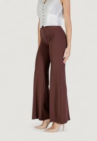 Pantaloni a zampa d'elefante marroni realizzati in tessuto elasticizzato, caratterizzati da una superficie liscia e una vita aderente. Abbinati a un top bianco senza maniche e tacchi nudi.