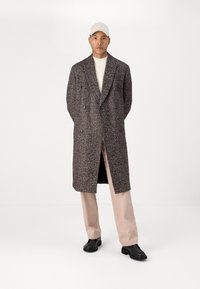 Abrigo de doble pecho, marrón y blanco moteado, hasta las rodillas, con solapas anchas; combinado con pantalones rosa claros y calzado negro.