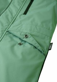 Jasnozielona wodoodporna tkanina o gładkiej fakturze, z kieszenią zapinaną na zatrzask i wszytą metką z logo, przedstawiona w pozycji złożonej.