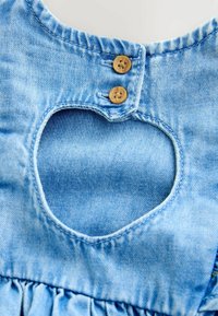 Robe en denim avec une découpe en forme de cœur dans le dos, couleur bleu clair, avec deux boutons en bois et des fronces au niveau de l'ourlet.