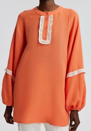 Dunkelhäutige Person trägt ein locker sitzendes, langärmliges orangefarbenes Oberteil mit weißer Spitzenverzierung an den Ärmeln und einer geknöpften Vorderleiste.