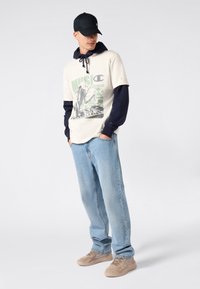 Mężczyzna w czapce na czarno, z warstwową beżową koszulką z grafiką na granatowym hoodie, w jasnoniebieskich dżinsach i beżowych sneakersach, stojący z rękami w kieszeniach.