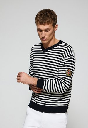 Pull rayé noir et blanc avec un col en V, des manches longues et des poignets côtelés. Présente un écusson sur la manche gauche.