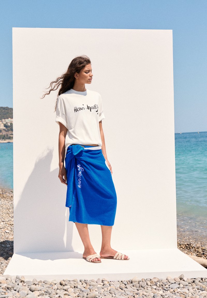Hvid t-shirt med sort tekst, blå indpakningsnederdel med knudedetalje og beige sandaler, sat mod en strand og minimalistisk baggrund.