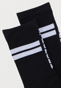 Μαύρες βαμβακερές κάλτσες με ραβδωτή υφή, που διαθέτουν δύο λευκές ρίγες και μία μαύρη ρίγα που διατρέχει οριζόντια κοντά στο επάνω μέρος.