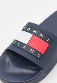 Tumesinine lipik sandaal paksu rihmaga, millel on mitmevärviline logo: punane, valge ja sinine, millel on kõrgendatud tähed "TOMMY JEANS". Sile tekstuur.