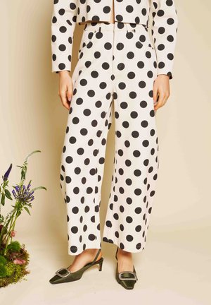 Kvinde iført hvide bukser med vide ben og cropped jakke med store sorte polkaprikker, kombineret med grønne kitten heel-sko med spænde.