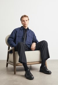 Tumesinine jakk kahe rinna taskuga, millele on sobitatud must lips ja püksid. Mustad nahkkingad beezis polsterdatud toolil.