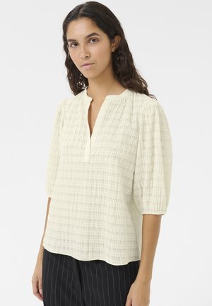 Blouse crème clair avec un motif texturé, manches mi-longues et encolure indentation. Le matériau semble léger et fluide. Associée à un pantalon rayé.