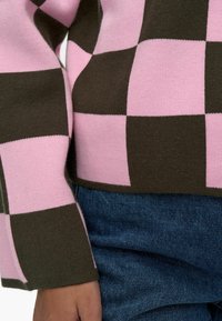 Maglione lavorato a maglia con un motivo a scacchi in rosa e marrone, con orlo e polsini a costine, indossato sopra un tessuto in denim.