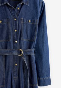 Combinaison en denim avec col, fermeture à boutons à l'avant, deux poches poitrine et une ceinture à la taille avec un anneau doré. Bleu foncé avec des accents dorés.