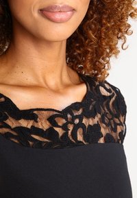 Schwarzes Kleid mit einem transparenten schwarzen Spitzenkragen und floralen Motiven. Glattes Gewebe darunter, das den Kontrast der Texturen hervorhebt.