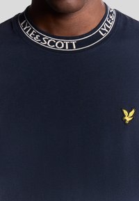Marineblå T-shirt med ribbet rund halsudskæring og "LYLE & SCOTT"-branding; inkluderer et lille gult ørner logo på brystet. Glat tekstur.