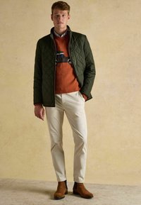 Mladý muž v olivovom prešívanom saku, hrdzavom svetri s grafikou auta, krémových nohaviciach a hnedých semišových topánkach stojí pri neutrálnej stene.