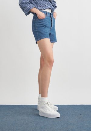 Personne debout les mains dans les poches portant un short en jean bleu, une chemise à carreaux bleue, des chaussettes blanches et des baskets montantes blanches.