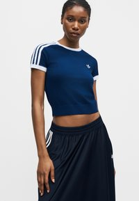 Femme portant un t-shirt court bleu marine avec des rayures blanches sur les manches et une jupe bleu marine avec des rayures blanches sur les poches.