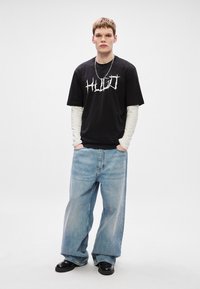 Μαύρο γραφικό μπλούζα T-shirt με λευκή γραφή, από πάνω από λευκό μακρύ μανίκι πουκάμισο. Φωτεινές μπλε φαρδιές τζιν παντελόνες και μαύρα παπούτσια ολοκληρώνουν την εμφάνιση.