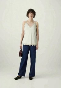 Lys grønne spencer med blonderkant kombinert med mørkeblå vidbukser, svarte loafers og en liten burgunder håndveske. Nøytral bakgrunn.