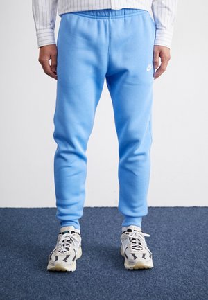 Mann trägt hellblaue Nike-Jogginghose und weiße Turnschuhe auf einem blau carpeted Boden vor einer weißen Wand.
