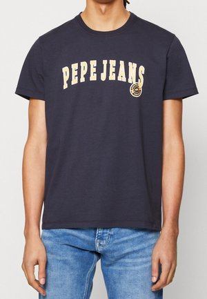 Osoba nosząca granatowy T-shirt z napisem „PEPE JEANS” dużymi beżowymi literami i małym okrągłym logo na piersi, w połączeniu z niebieskimi dżinsami.