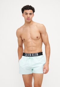 Minta zelena kopalna kratka hlača z črnim pasom, na katerem je logotip "CALVIN KLEIN"; vsebuje stranske žepke in vrvico za prilagajanje.