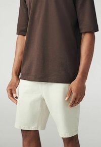T-shirt en coton marron avec des manches roulées, associ�é à un short en denim vert clair ayant une coupe droite et un ourlet retroussé. Aucune quincaillerie visible.