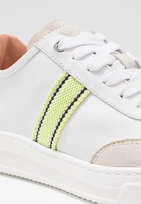 Baskets en cuir blanc avec une bande texturée jaune néon et noire, des accents en daim gris, et des lacets blancs sur une semelle épaisse blanche.