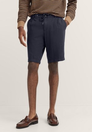 Marineblaue Shorts aus leichtem Stoff mit einem Kordelzugbund, ausgestattet mit Falten und umgeschlagenen Säumen, kombiniert mit braunen Lederloafers.
